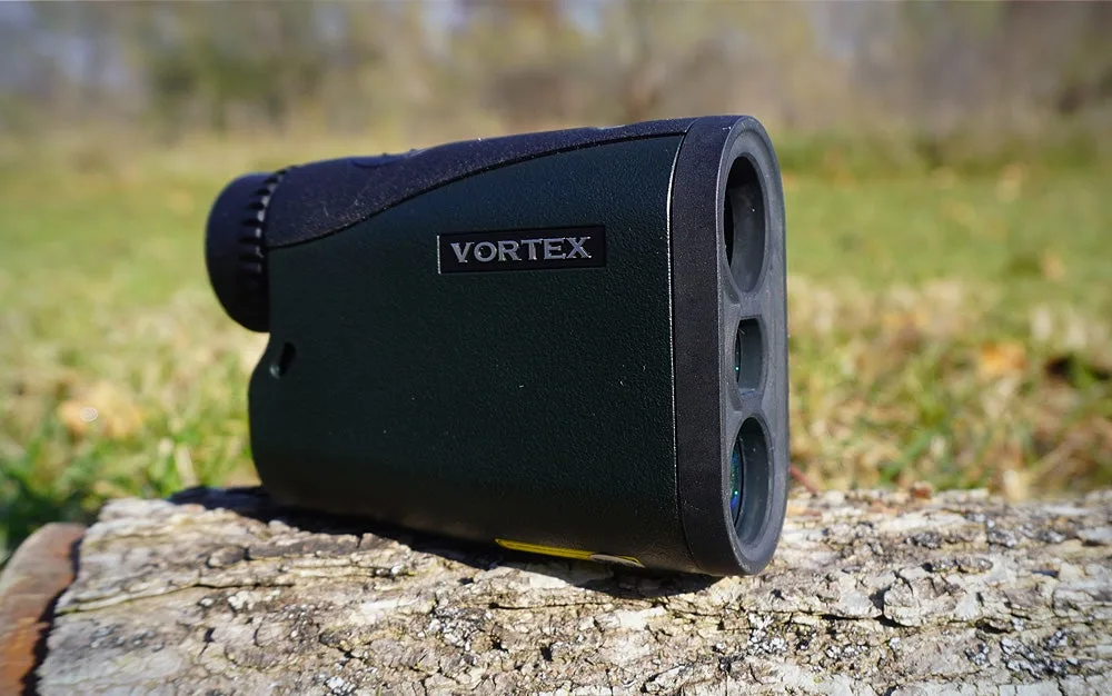 A Vortex Crossfire HD 1400 rangefinder sitting on a gray log in a grassy lawn.