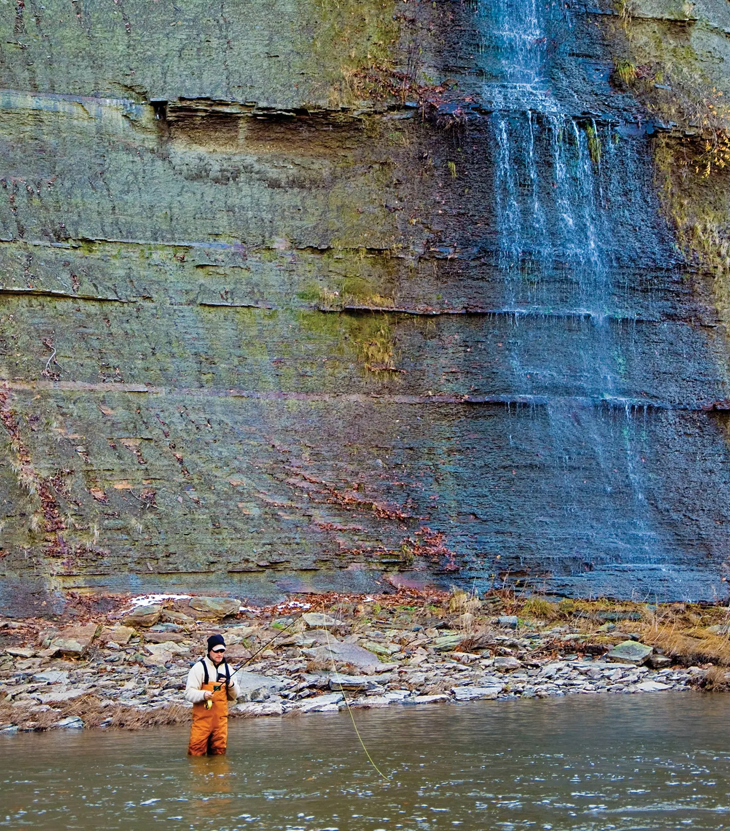 Anglers holding fly rod stands in knee-deep river, waterfall behind him has left bright blue deposits on rock.