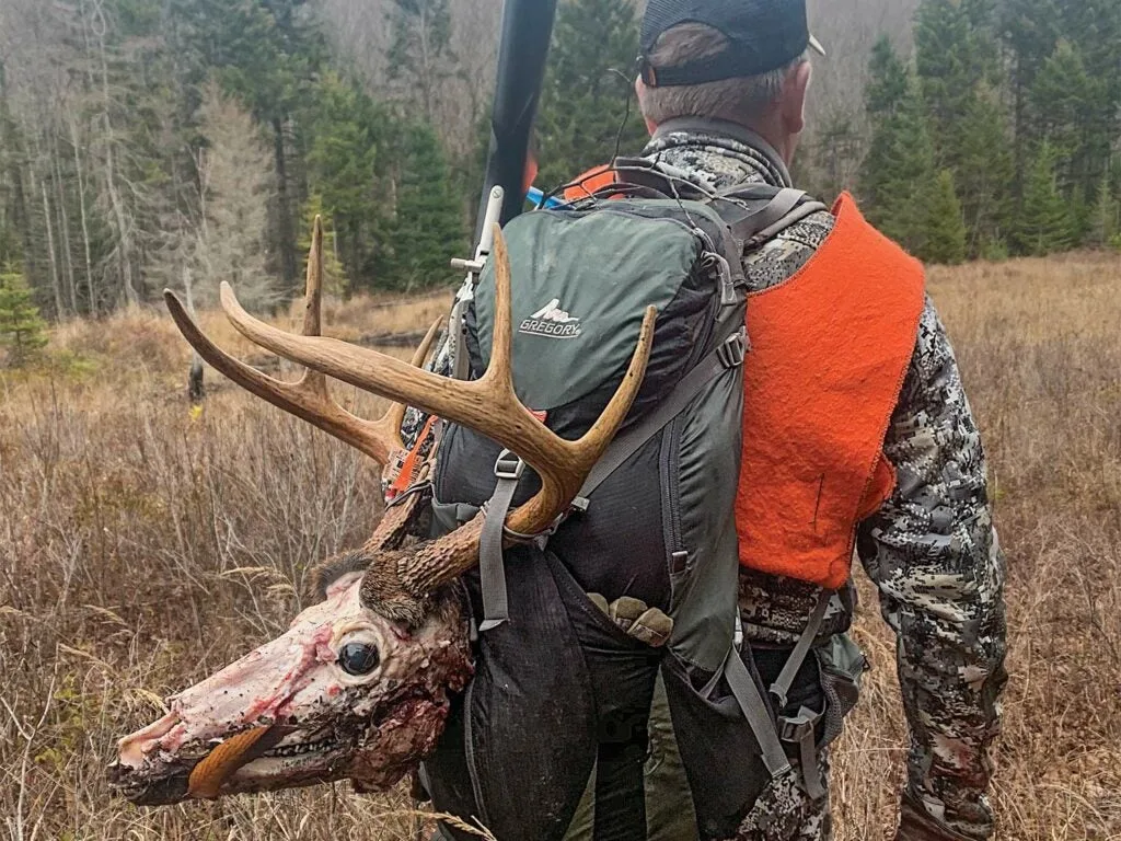 A hunter walks through a field with a deer head strapped to his back.