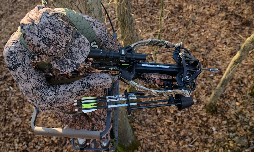 A hunter shoots a crossbow from a tree stand in the woods.