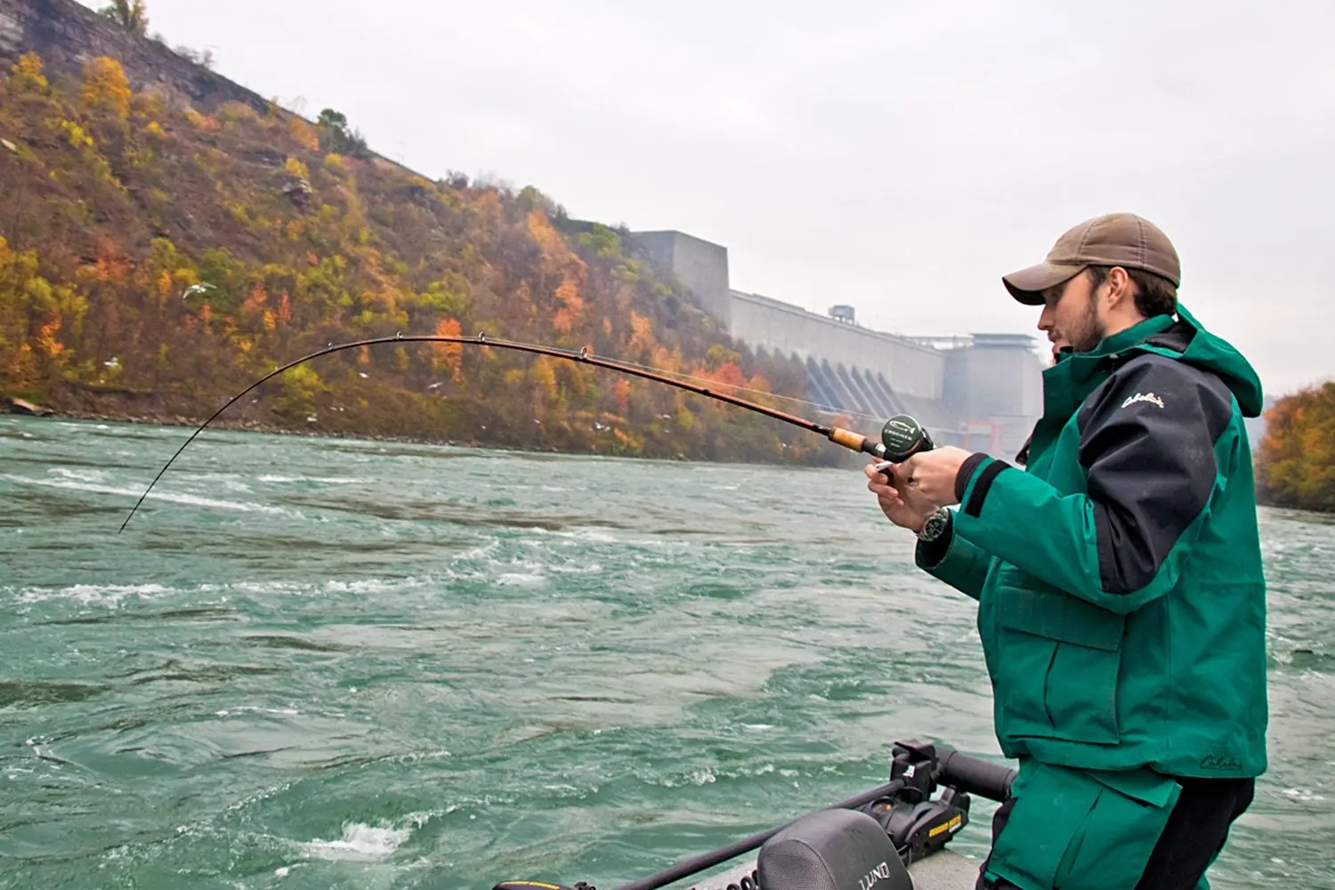 Angler dressed in waterproof clothing and standing on boat holds bent rod.