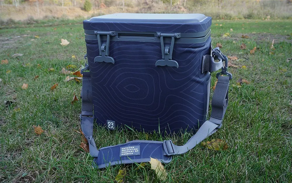 The blue and gray Orca Wanderer 24 cooler with shoulder strap sitting on green grass.