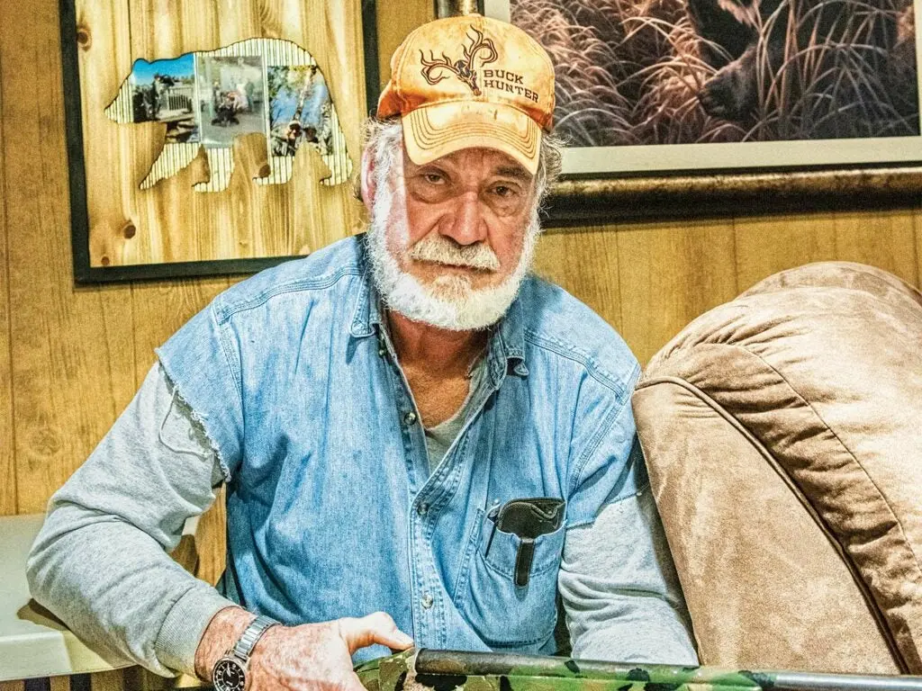 An elderly man holds a camo shotgun and looks at the camera while seated in a char in a bear camp house.