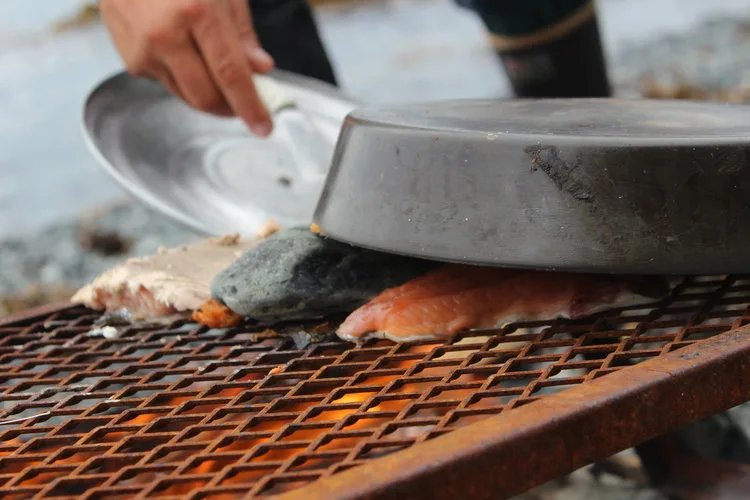 Wild salmon being cooked over an open flame on the shore of a river