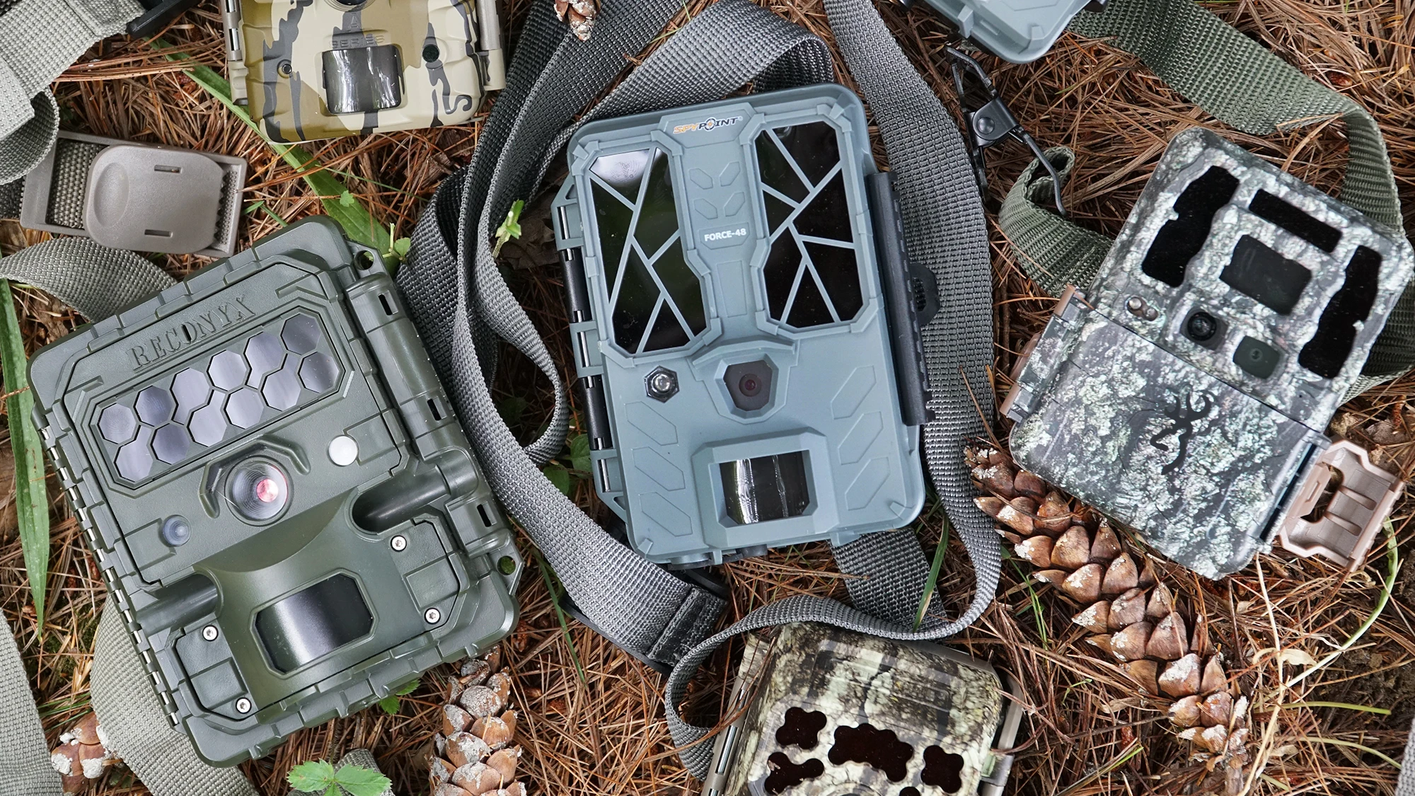 A selection of new conventional trail cameras lying in pine duff near pine cones.