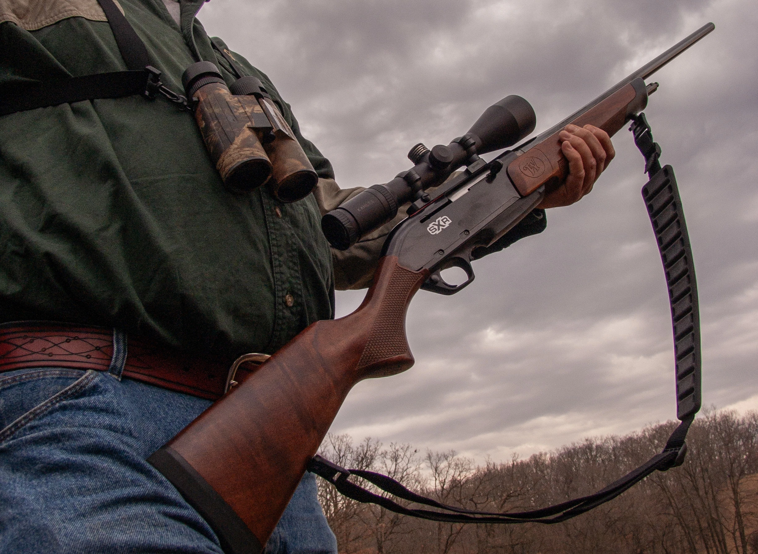 A shooter holds a scoped rifle that has a carry strap. 