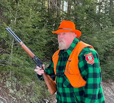 A hunter wearing blaze orange and a green-checked jacket walk the woods with a lever-action rifle. 