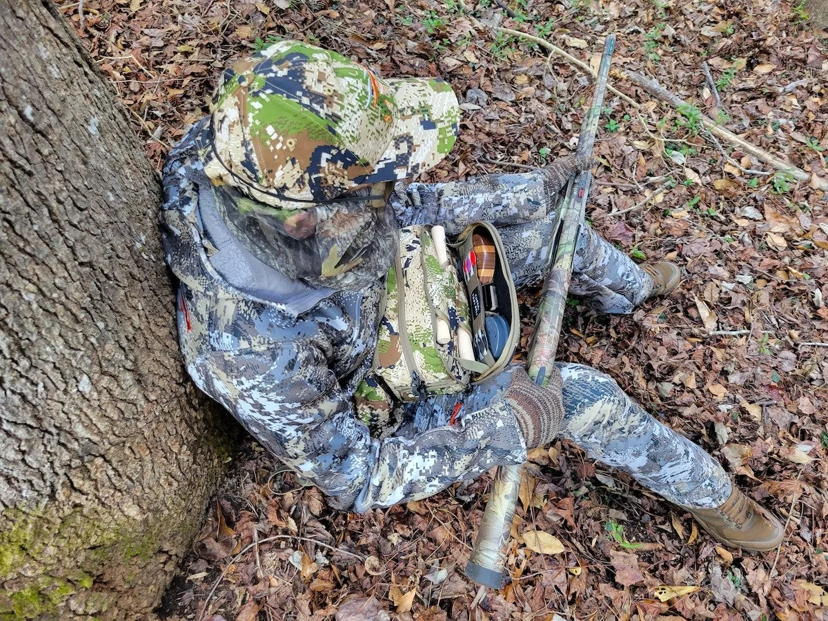 Female turkey hunter sitting against tree with shotgun and Sitka tool belt