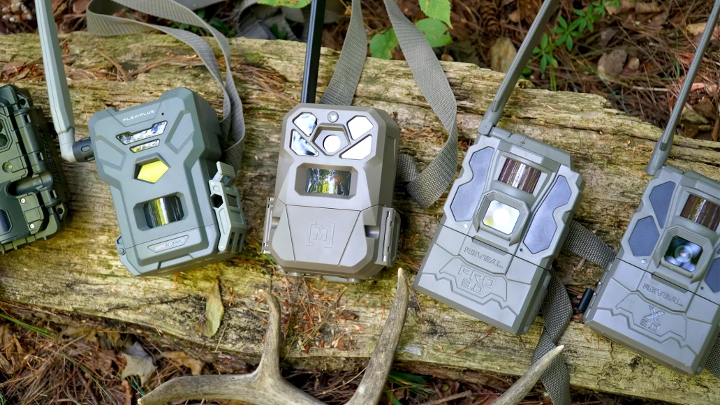 Five cellular trail cameras rest on a log in the woods, with part of a deer antler showing.