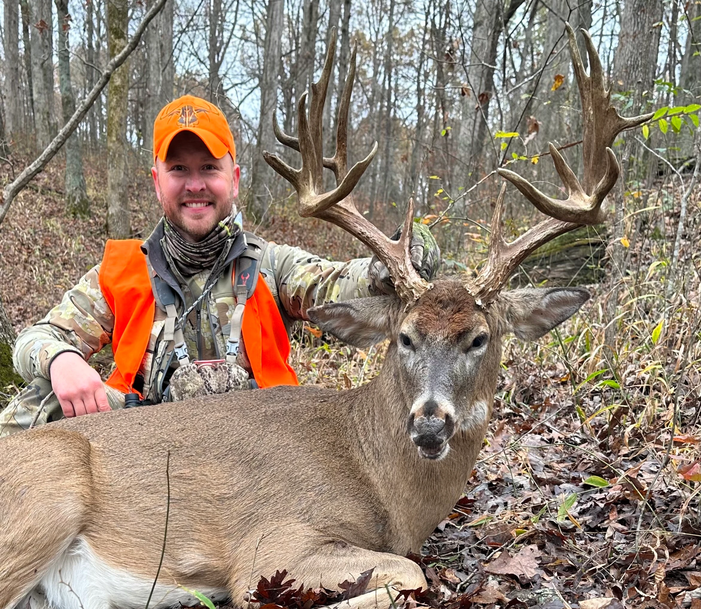 A hunter in orange sits on the ground in the woods and poses with a trophy whitetail buck.