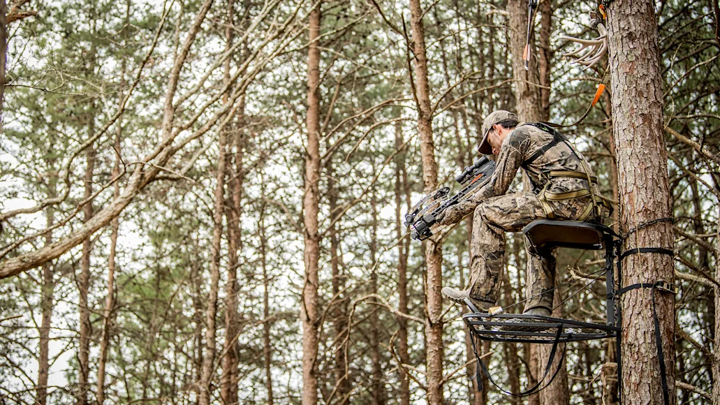 A hunter shoots a crossbow from high in a tree stand, with open woods in the background.