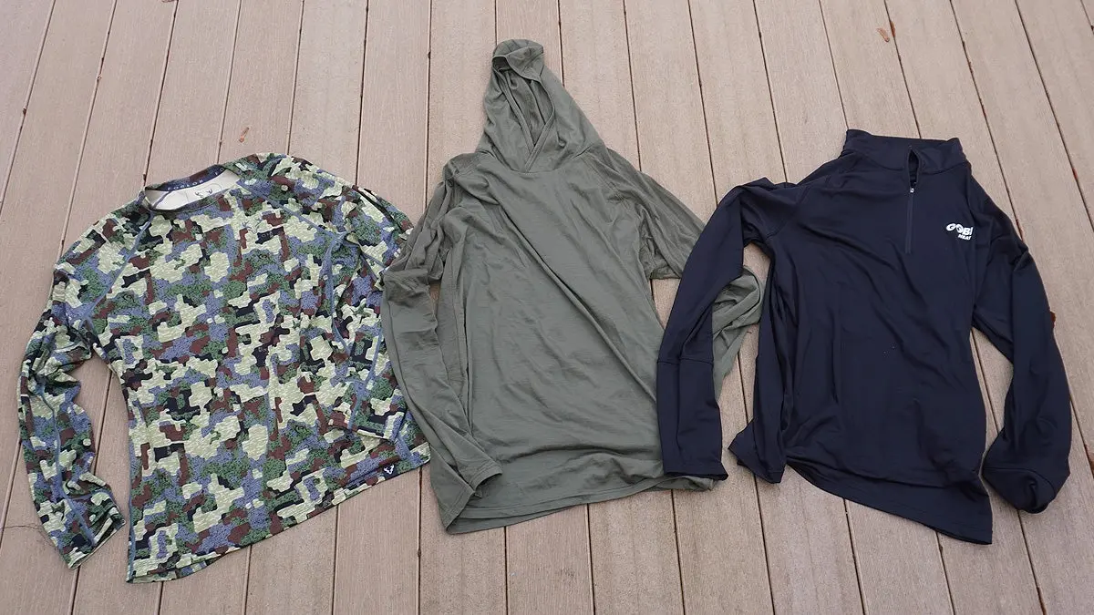 Three base layer shirts for hunting in camo, green, and black sitting on a tan deck.