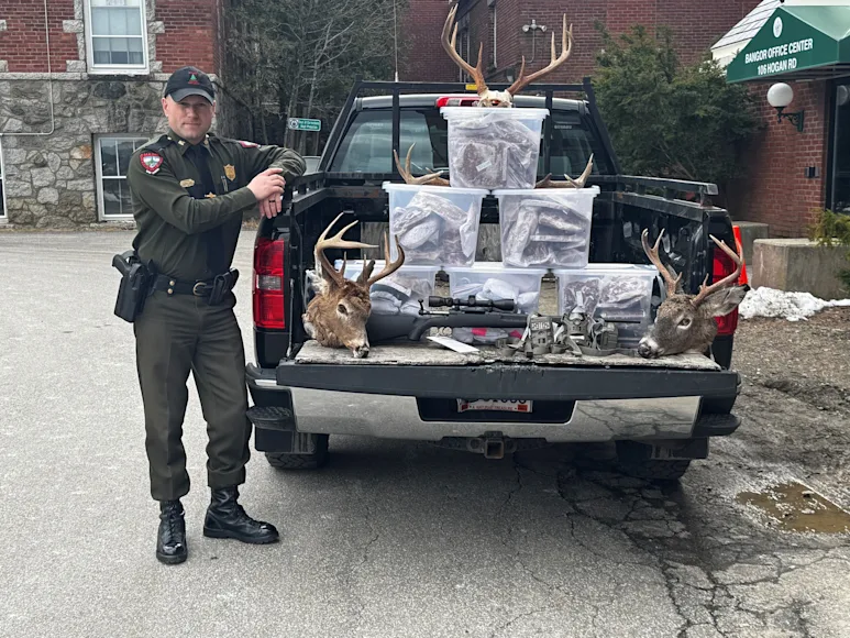 A Maine game warden poses with confiscated deer heads and rifles.