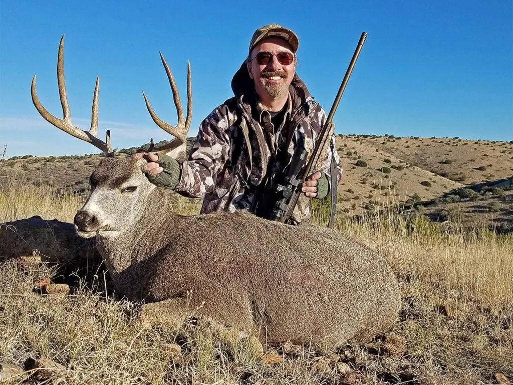 A hunter holding a ruger american rifle while kneeling behind a deer.