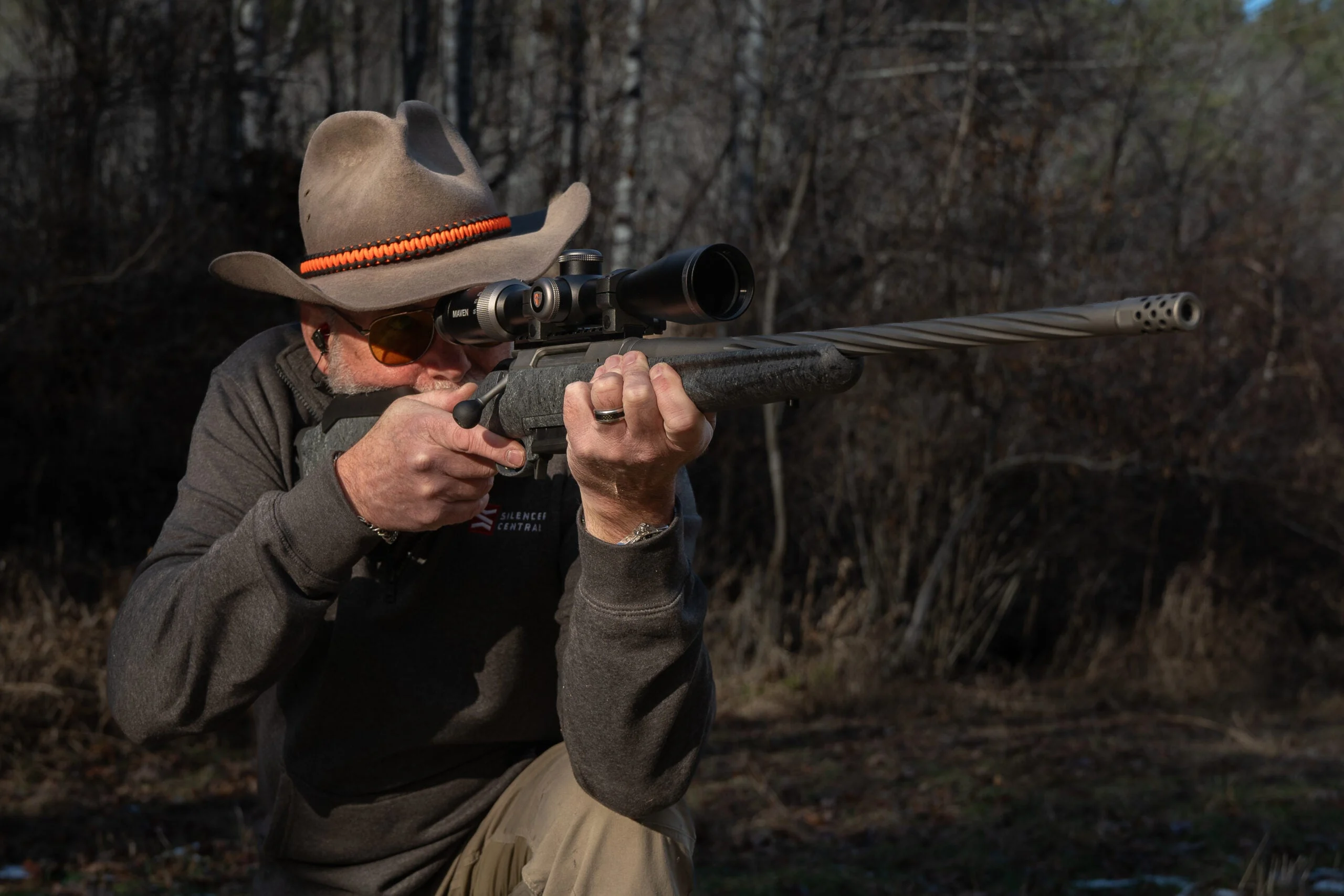 Shooter fires rifle from a kneeling position with woods in background