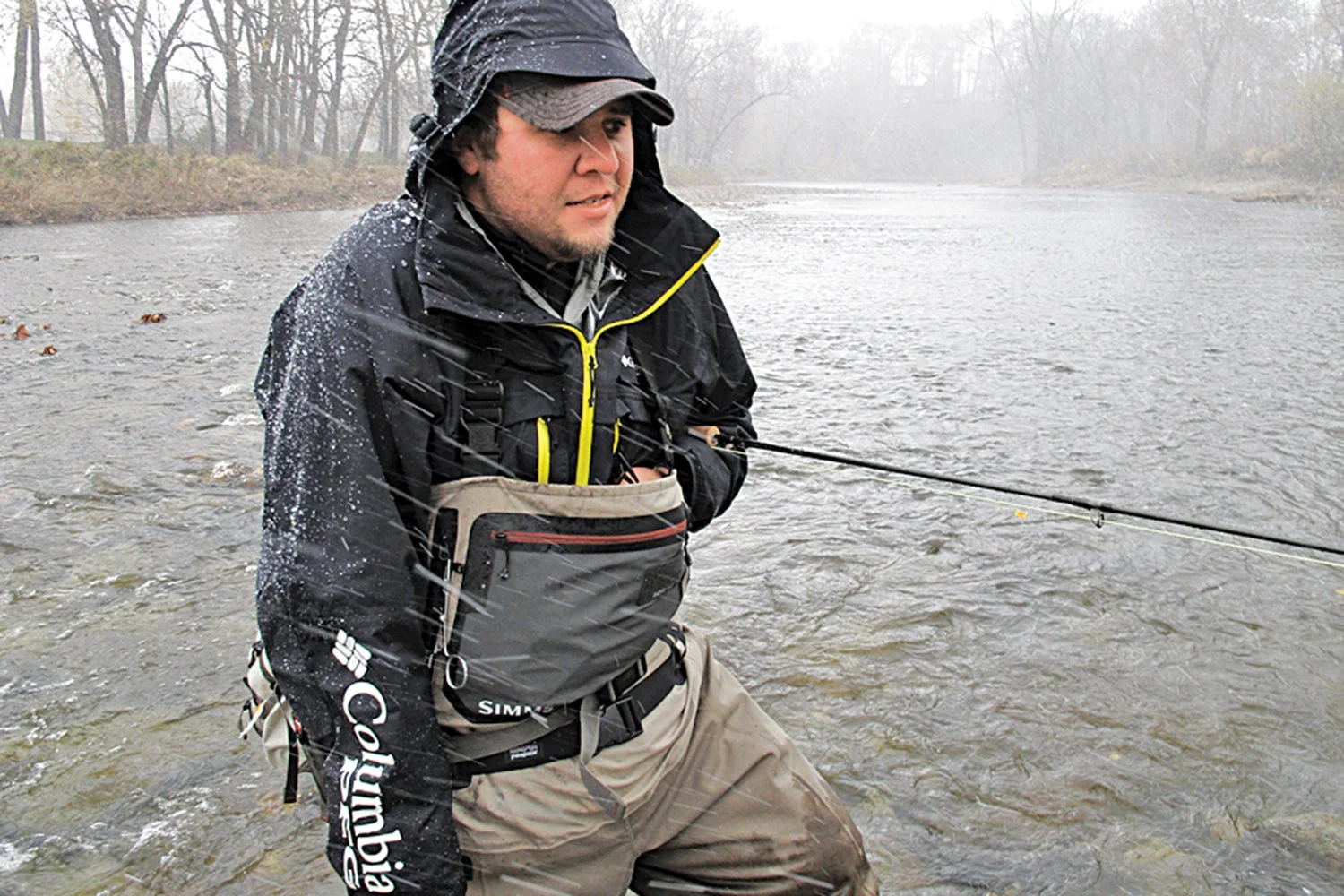 cold angler stands in knee-deep river with hands stuffed in pocket and rod tucked under arm and snow falls around him