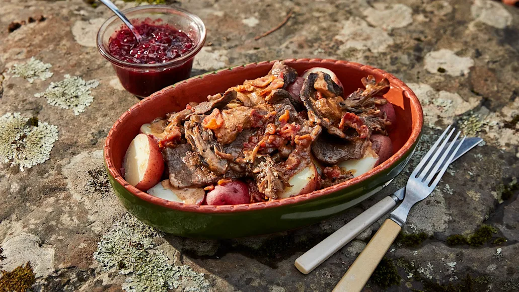 bowl of venison with potatoes and bacon sits on lichen-covered rock along with fork, knife, and small condiment bowl containing red berries