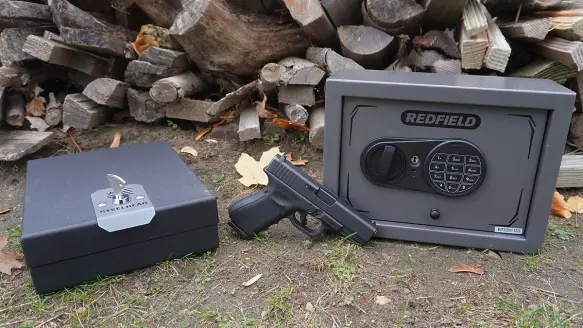 Two black and grey handgun safes and a Glock 19 handgun sitting on the ground in front of pile of firewood.