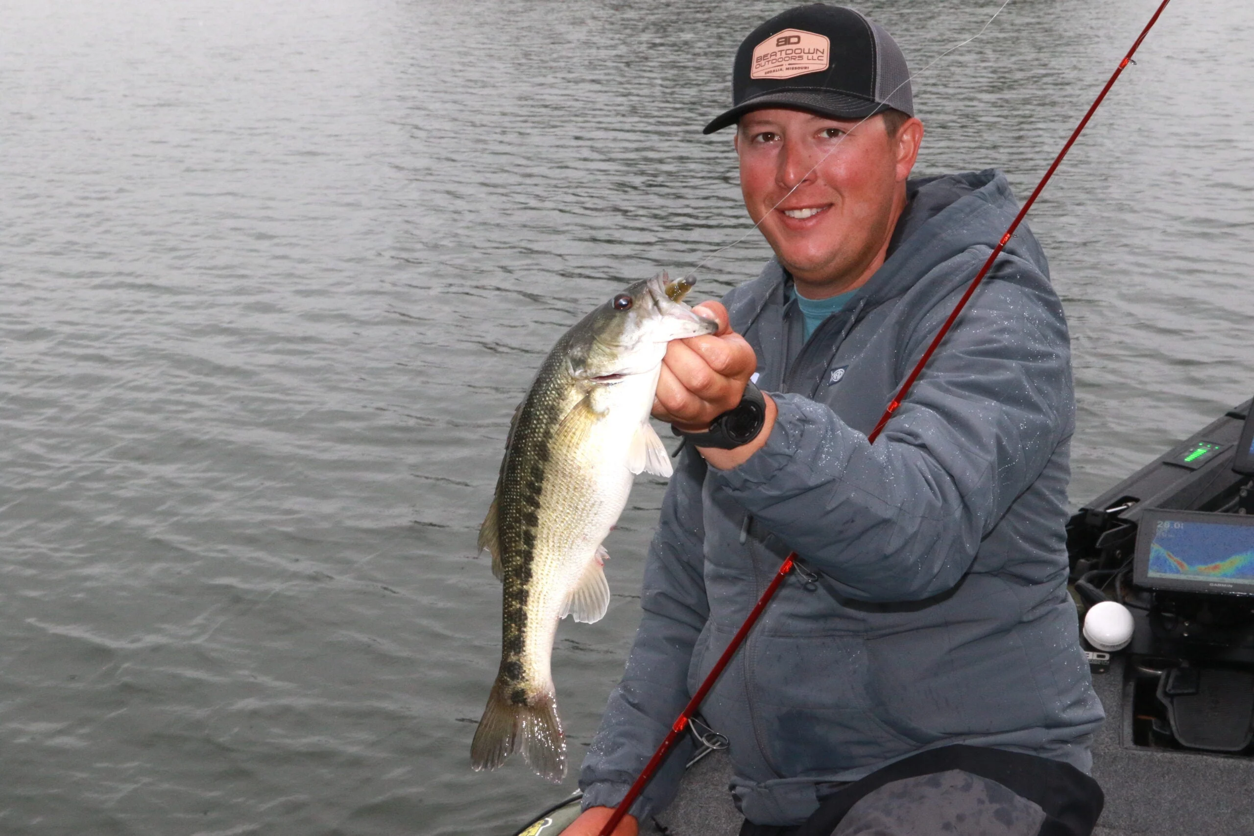 An angler in a boat shows off a bass caught on a jighead minnow, with lake in the background.