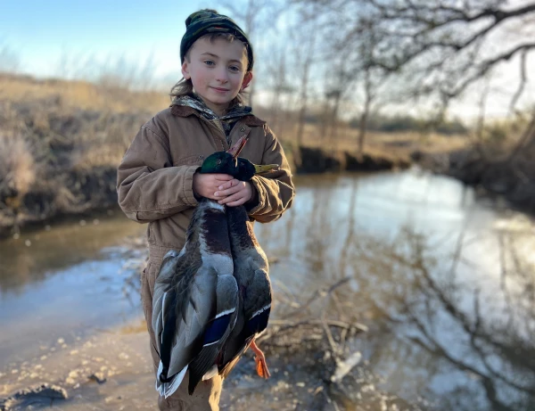 boy holds two ducks and stands in front of pond in lightly wooded area