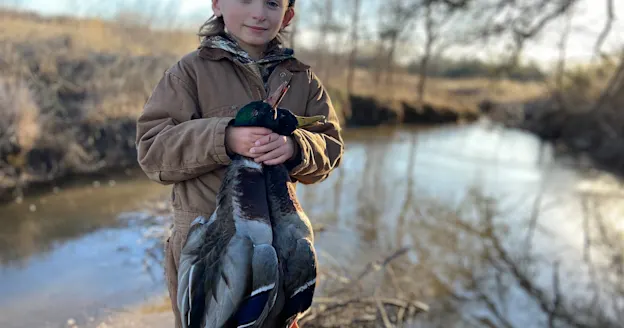 boy holds two ducks and stands in front of pond in lightly wooded area