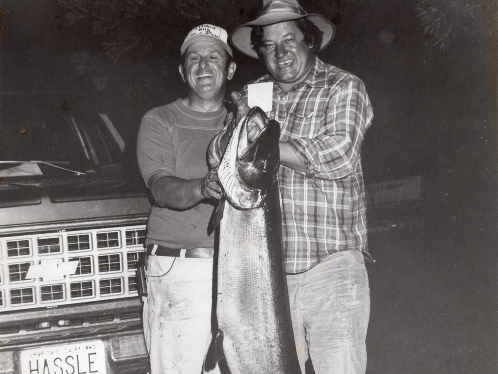 A black and white photo of two men holding up a large muskie fish.