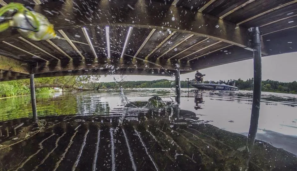 A bass angler skips a bait under a dock, with the bait splashing in the foreground.