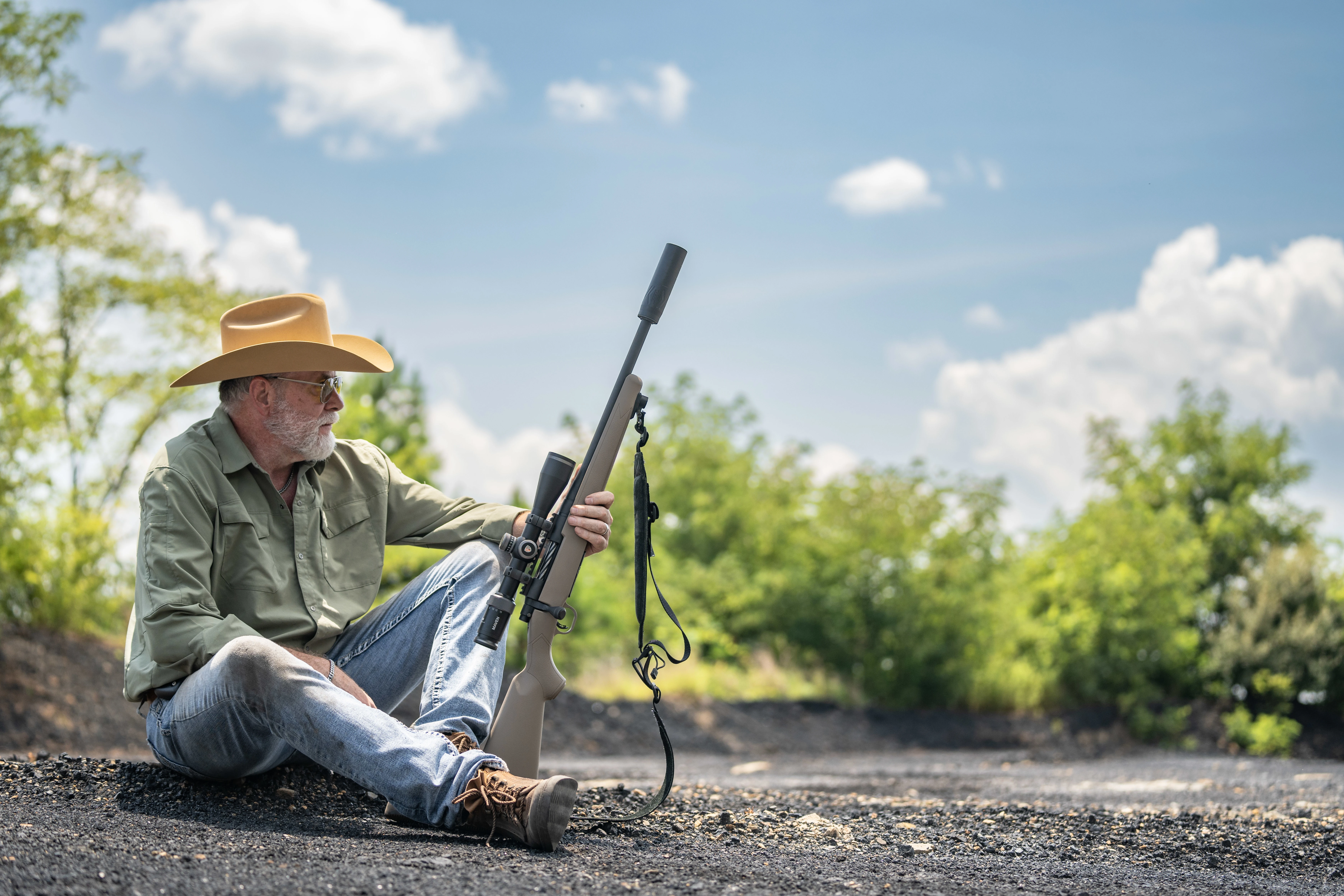 A man in a straw hat and green shirt sits on the ground holding a hunting rifle at a shooting range