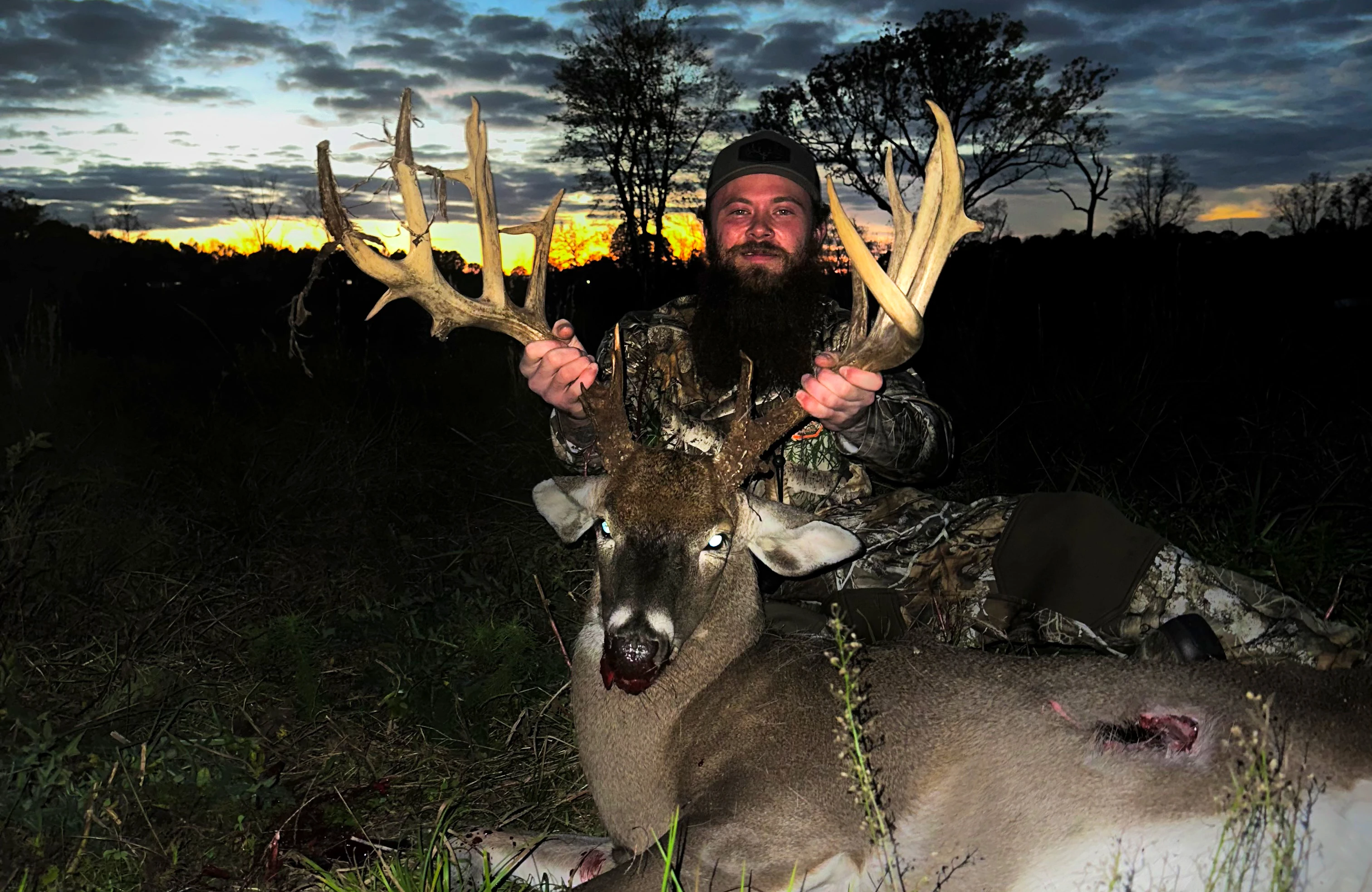 A hunter in a field poses with a big whitetail buck at dusk. 