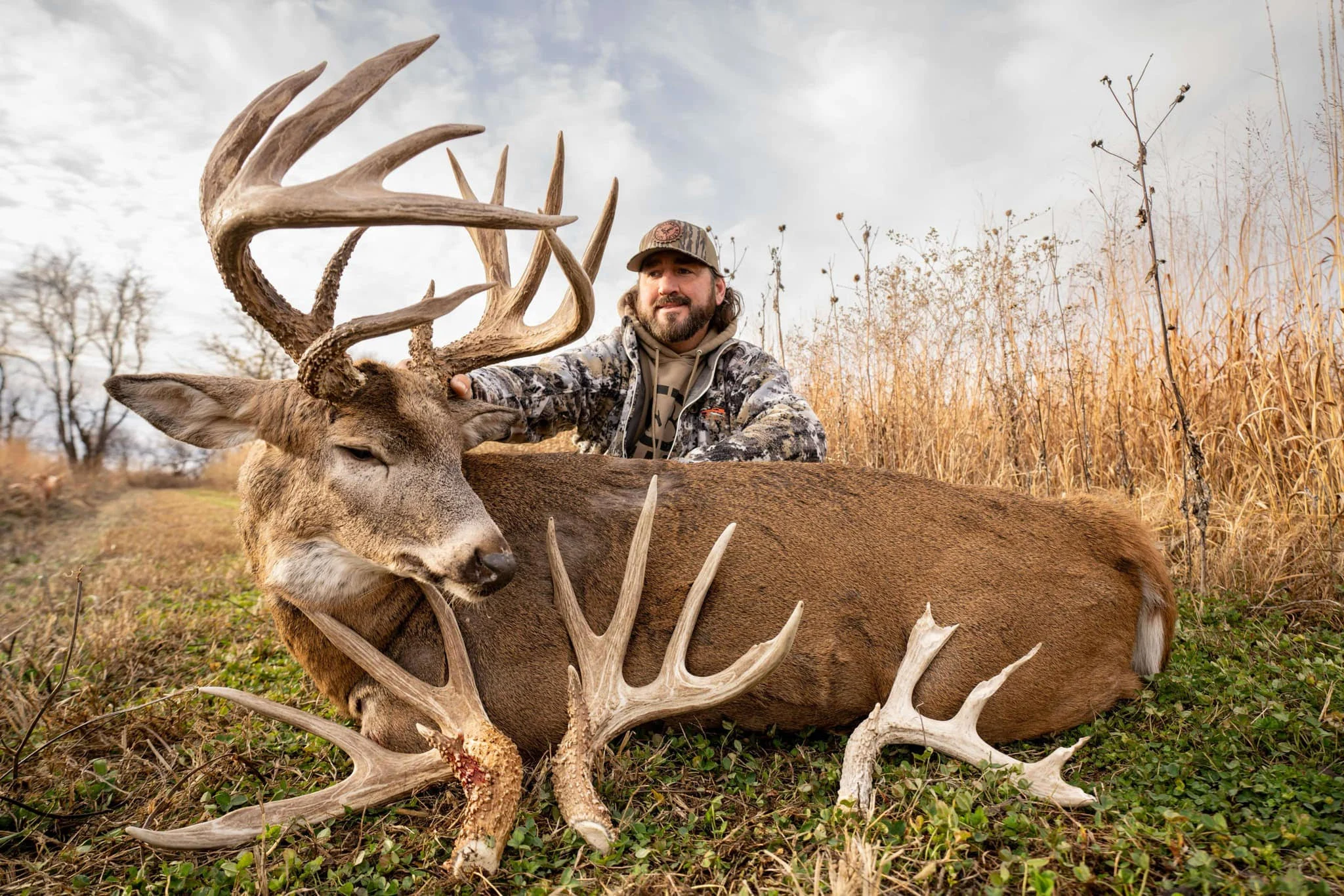 A hunter sits on the ground in a field and poses with a huge whitetail buck, the deer's sheds visible in the foreground. 