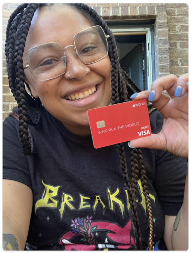 Young woman holding up the orange Sequin Visa® Debit Card, with writing on the card being "Who Run the World"