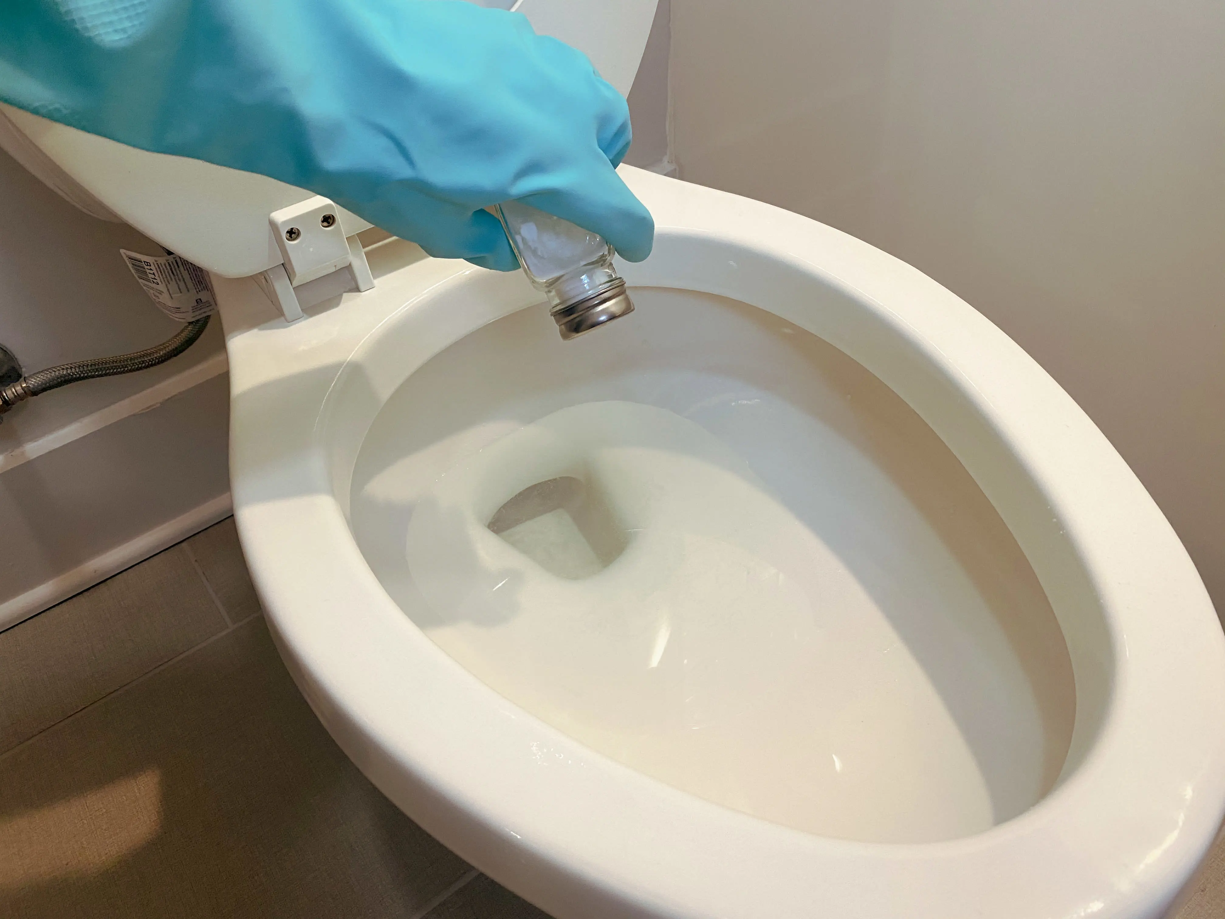 A person wearing gloves shakes baking soda into a toilet with a salt shaker