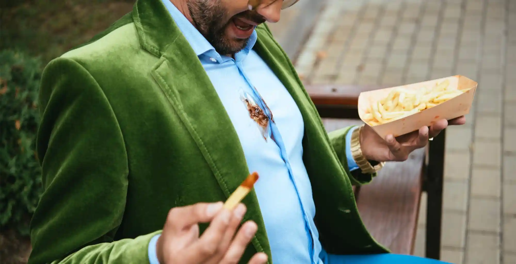 Un hombre, vestido de forma elegante con una mancha de kétchup en su camisa, sostiene unas papas fritas sentado en un banco en la calle