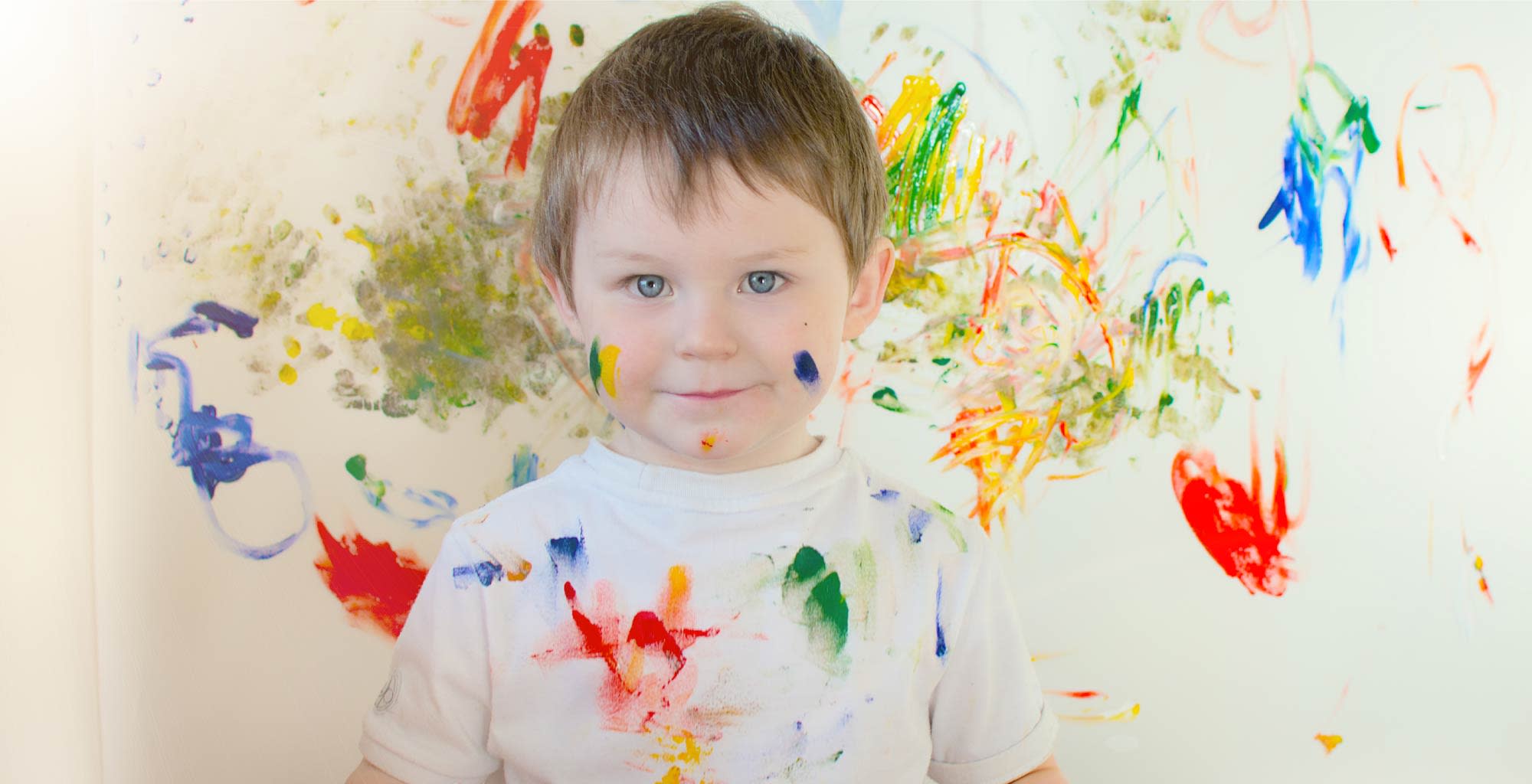 Un niño manchado de pintura parado junto a una pared llena de garabatos.