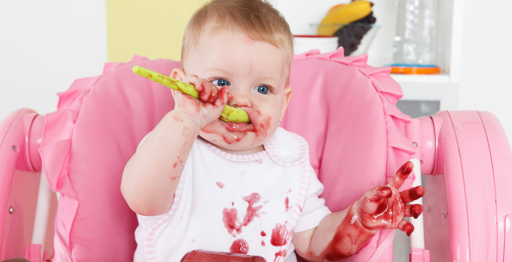 Una bebé sentada en su silla comiendo un postre mientas se mancha.