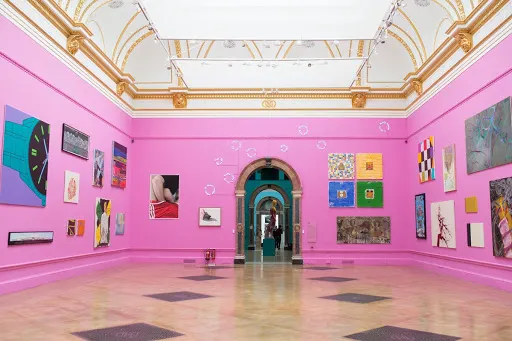 A brightly lit art gallery with pink walls displays various colorful paintings and artworks. The ornate ceiling has gold detailing, and a central arched doorway leads to another room.