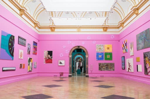 A brightly lit art gallery with pink walls displays various colorful paintings and artworks. The ornate ceiling has gold detailing, and a central arched doorway leads to another room.