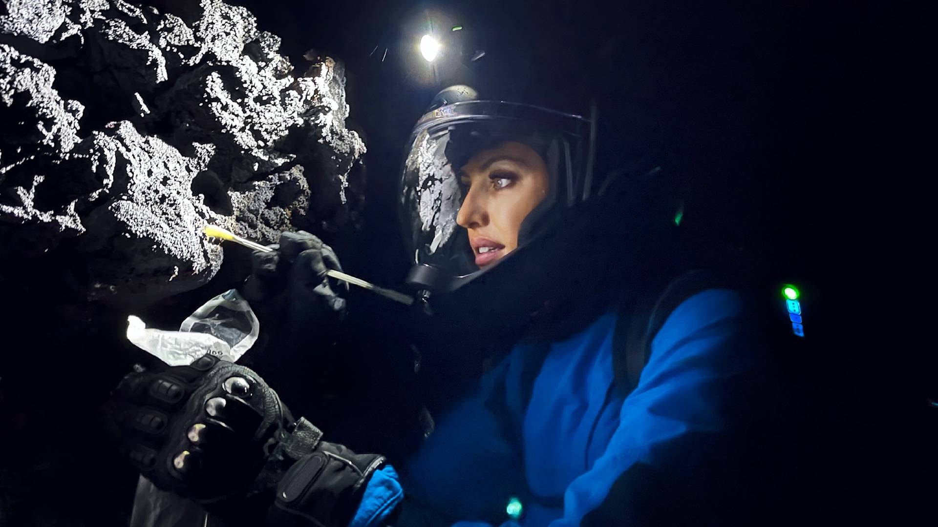 A person in a helmet and protective gear collects a rock sample with a tool inside a dark cave, illuminated by a headlamp.
