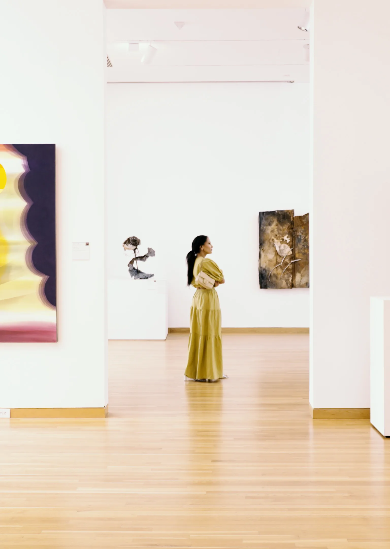 A woman in a long yellow dress stands in a bright art gallery, looking at various artworks on white walls. The floor is wooden, and several abstract pieces hang or are displayed around her.