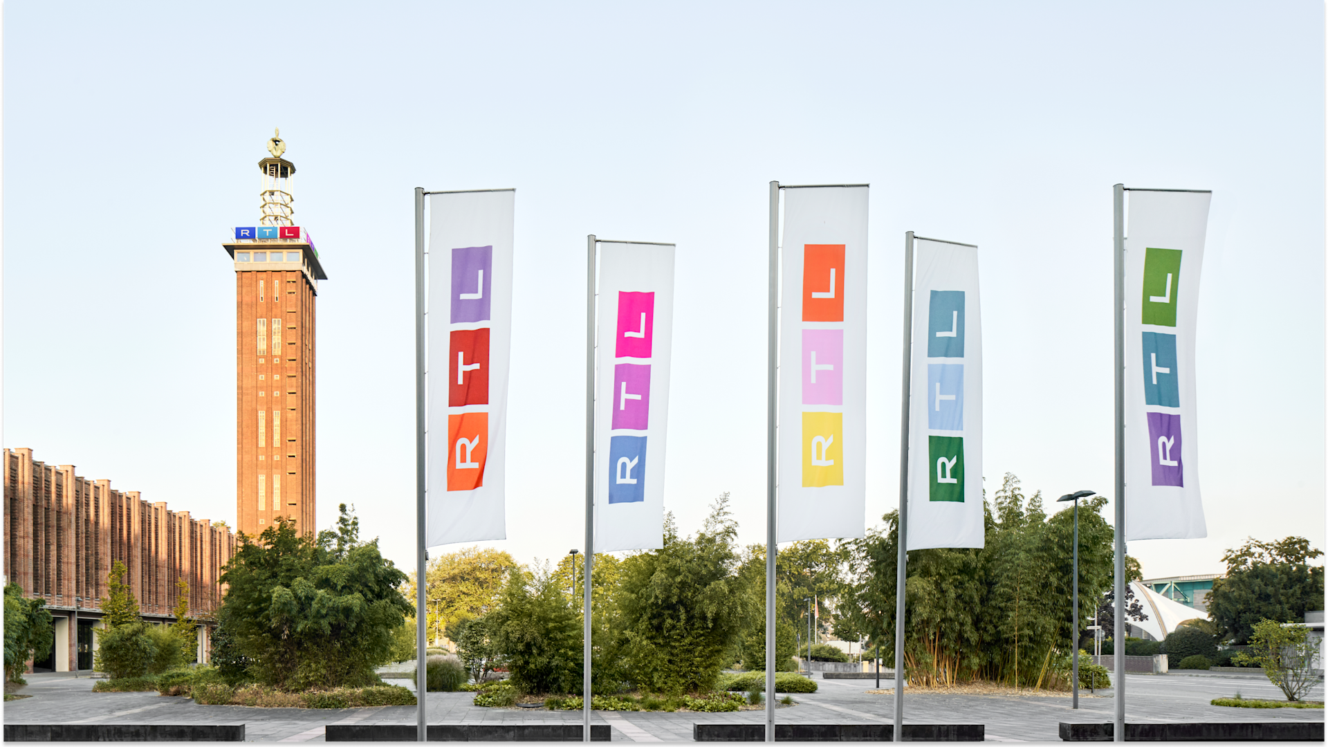 A row of five RTL flags in various colors stands in front of a brick tower with a clock and antenna, surrounded by greenery under a clear sky.