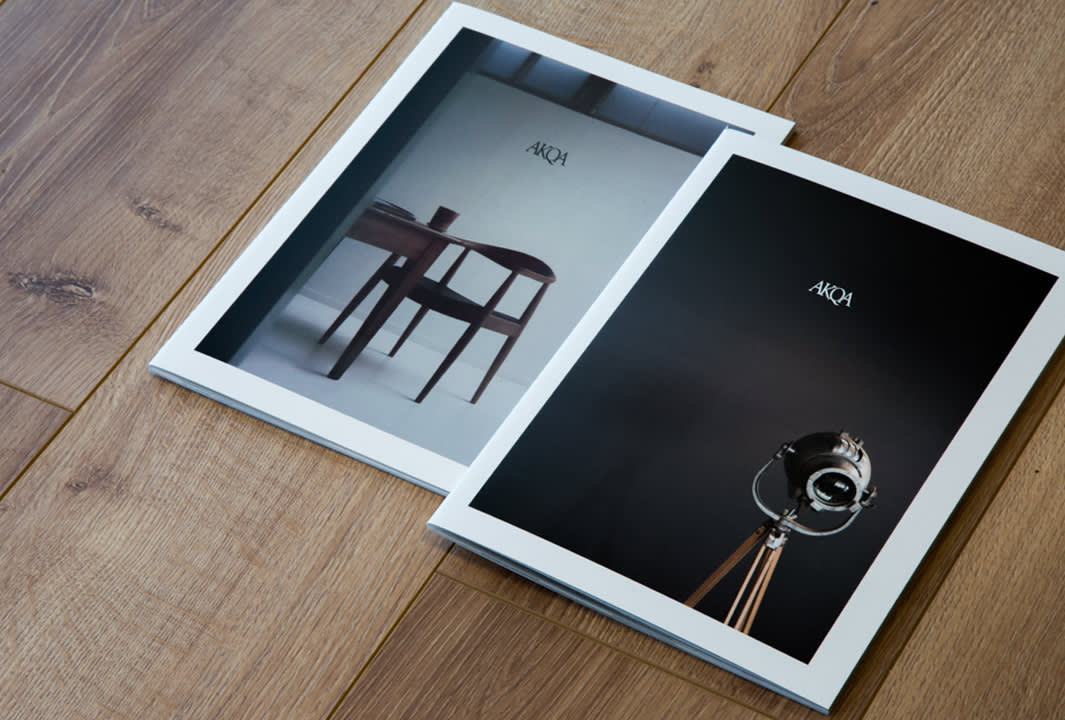 Two magazines with minimalist covers lie on a wooden floor; one shows a chair and table, the other features a tripod lamp. Both covers have AKQA written at the top.