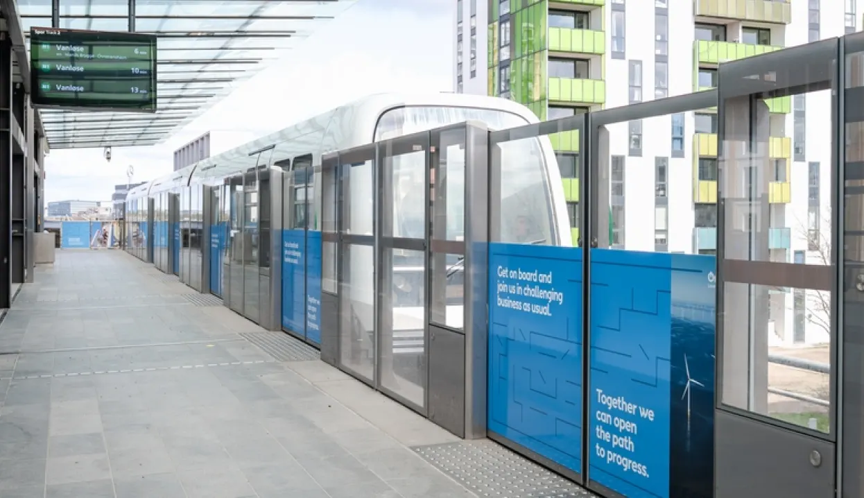 A modern light rail train stops at a clean, open-air station with glass barriers. Tall, colorful buildings are in the background. Blue panels on the barriers display messages about progress and sustainability.