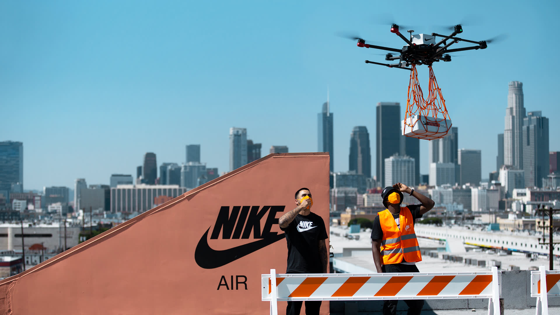 A large drone delivers a package above a rooftop with a Nike logo, as two workers in safety vests and helmets look on. The city skyline is visible in the background under a clear blue sky.