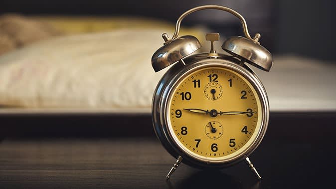 A vintage yellow alarm clock with two bells sits on a dark wooden bedside table, with a blurry pillow and bed in the background.
