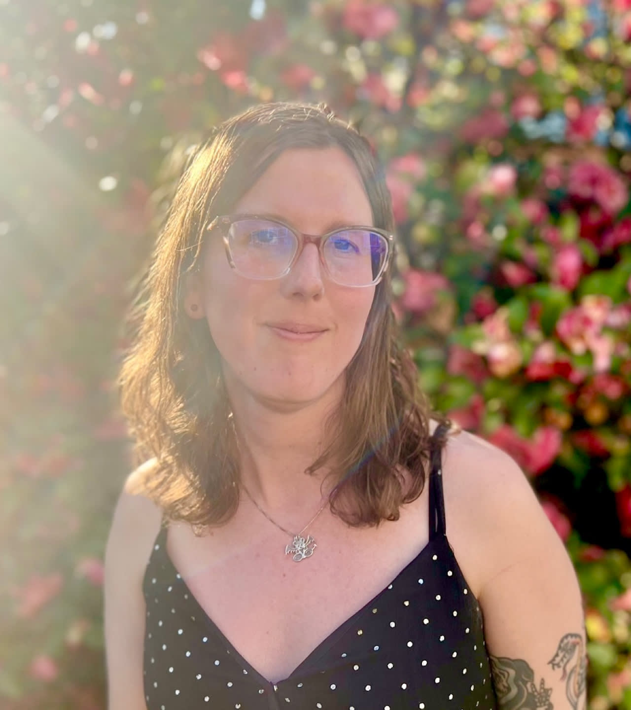 A person with shoulder-length brown hair and glasses stands outdoors in sunlight, wearing a black polka-dot top. Pink flowers and green leaves are in the blurred background, and a tattoo is visible on their upper arm.