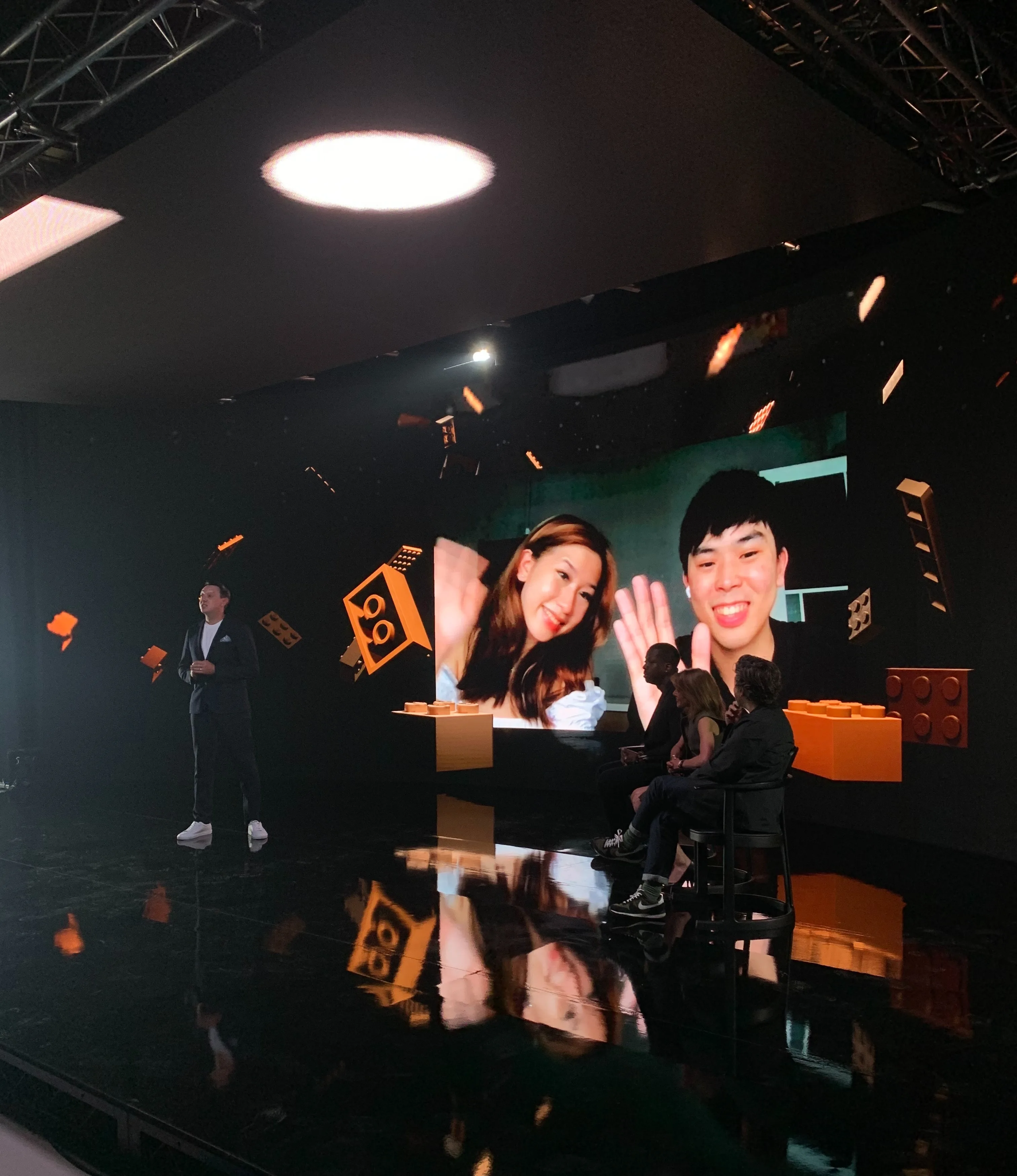 A man stands on stage next to a large screen showing two people smiling and waving. Three seated individuals face the screen. Digital LEGO bricks are displayed in the background. The setting appears to be a modern event or presentation.