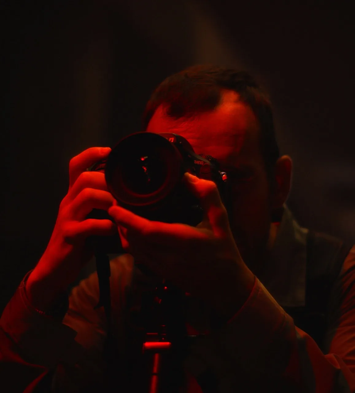 A person is holding a camera up to their face, preparing to take a photo. The scene is dimly lit with dramatic red lighting, casting strong shadows and highlighting the photographers hands and facial outline.