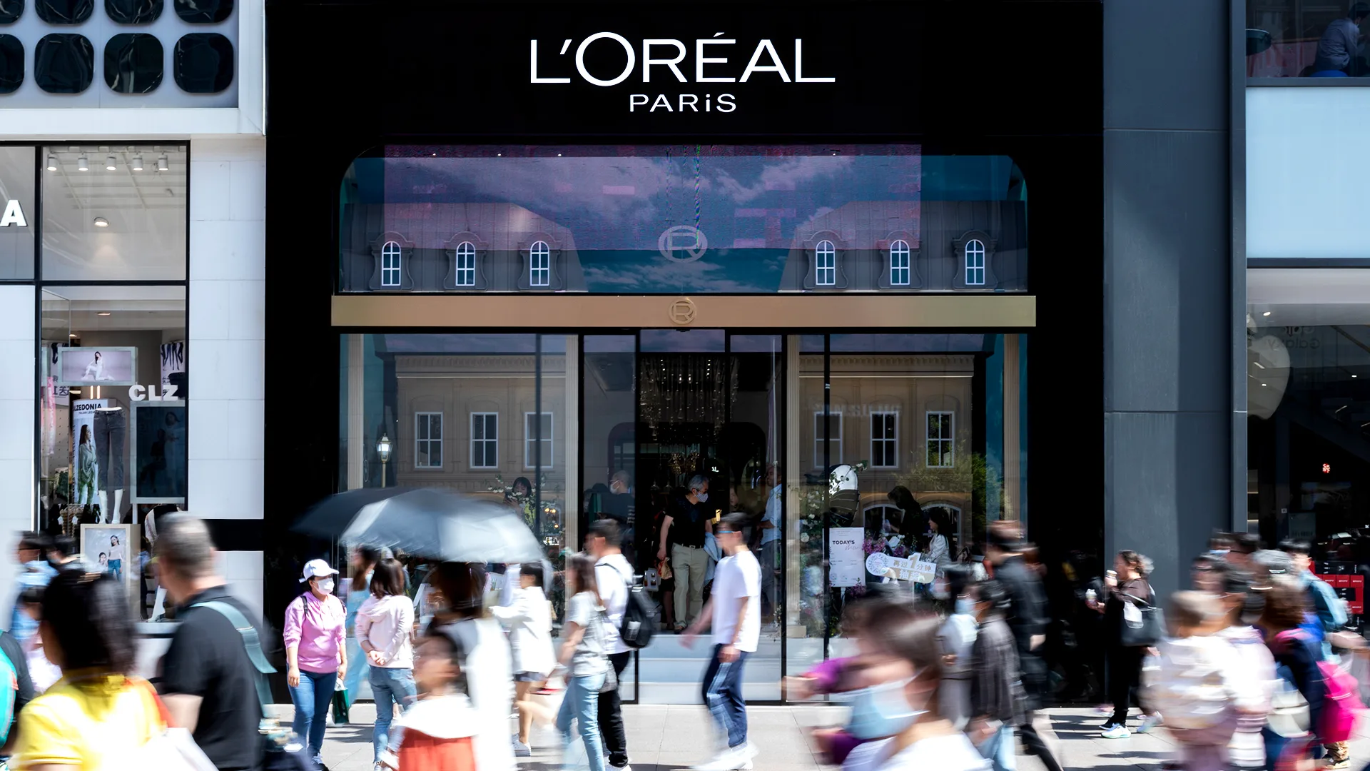 Crowds of people walk past a modern LOréal Paris store with large glass windows and a black sign above the entrance on a busy city street.