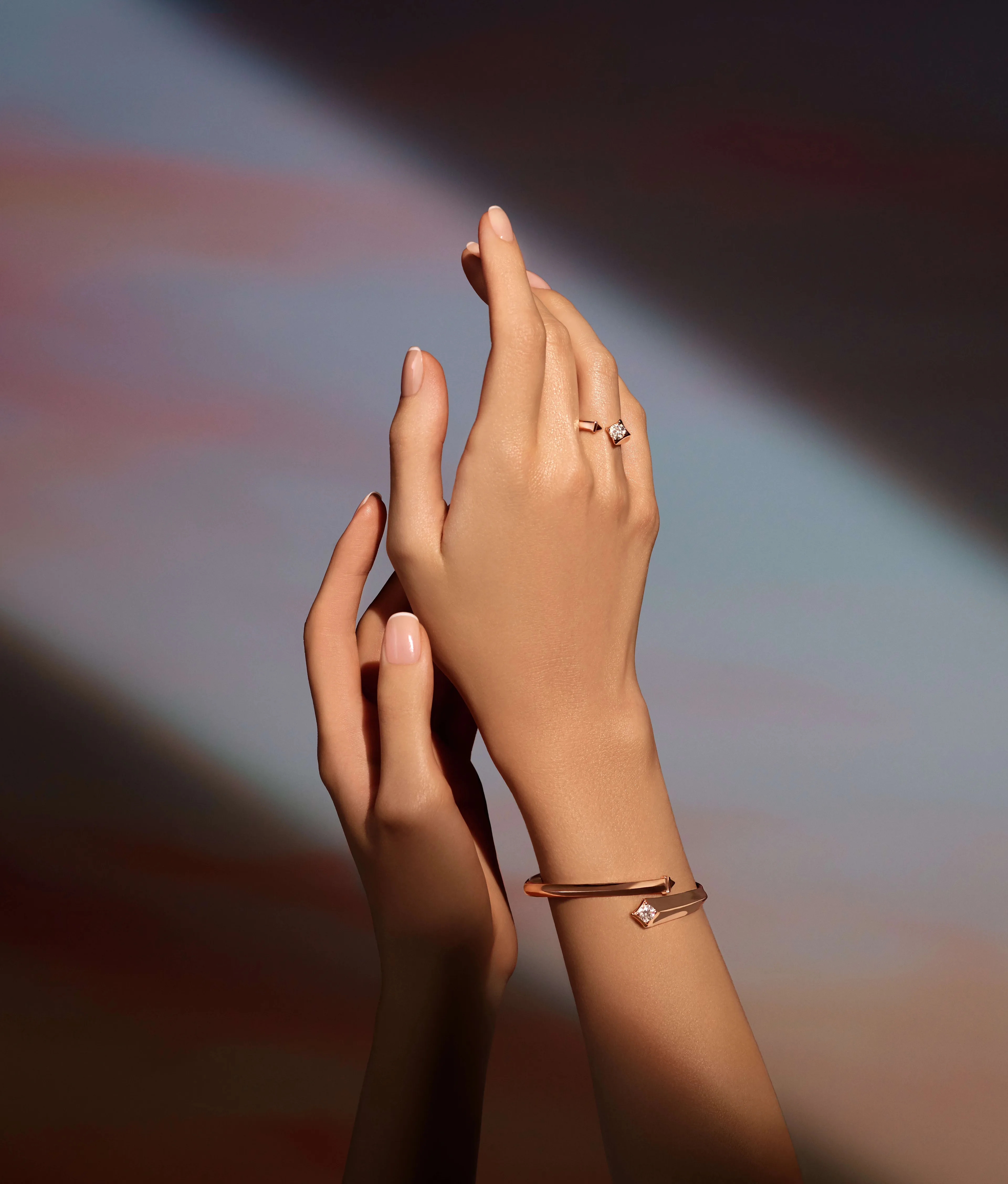 Two elegantly manicured hands with nude nail polish are posed gracefully. One hand wears a gold ring with a small stone, and the other wrist has a delicate gold bracelet with a gem. The background is softly blurred.
