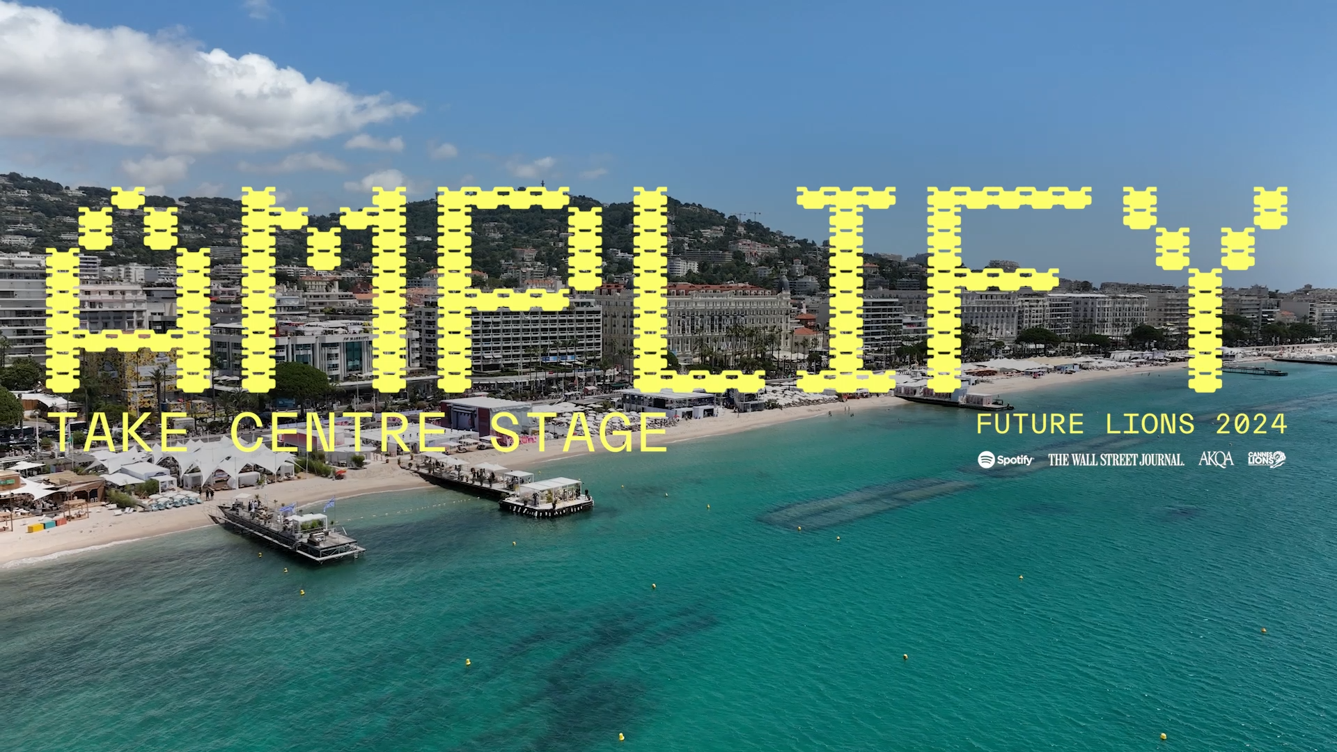 Aerial view of a sunny beach with turquoise water and piers, overlaid with yellow pixelated text reading AMPLIFY, and smaller text underneath saying, Take Centre Stage, Future Lions 2024.
